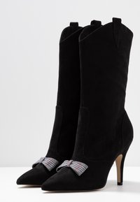 Bottines en suède noir avec des bouts pointus et un talon haut. Présentent un nœud décoratif en plaid gris sur le devant et des découpes sur les côtés.