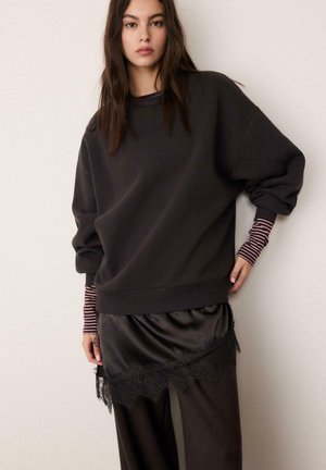 Jeune femme aux longs cheveux bruns portant un sweat-shirt noir surdimensionné, des manches longues rayées, une jupe noire bordée de dentelle sur un pantalon noir, fond neutre.