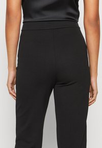 Pantalon taille haute noir en tissu lisse et extensible. Présente une coupe ajustée, des coutures latérales et une texture subtile. Aucun élément métallique visible.