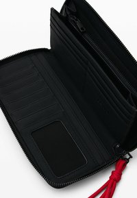 Portafoglio in pelle nera con più fessure per carte, una tasca con cerniera e un tirazip in tessuto rosso. Texture liscia e design minimalista.