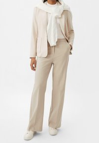Mujer vistiendo un blazer beige, pantalones anchos a juego, zapatillas blancas y un suéter blanco sobre los hombros.