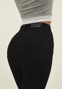 Vaqueros negros de talle alto, con una textura suave, costuras sutiles y dos bolsillos traseros. Etiqueta del logo visible en la cintura.