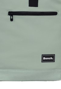 Sac à dos vert clair avec une finition texturée. Équipé d'une poche zippée noire et d'un logo « Bench. » sur une étiquette noire rectangulaire.