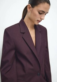 Bordowa marynarka o dopasowanym kroju, z klapami z wycięciem, zapinana na jeden guzik, z kieszenią na piersi. Wykonana z teksturowanego materiału.