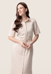 Robe chemise beige à manches courtes en tissu lisse, avec un devant à boutons, une ceinture avec boucle et un détail de bas plissé.