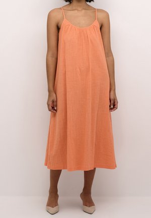 Vrouw draagt een losse, mouwloze midi-jurk in perzikkleur met dunne bandjes en beige schoenen met spits toelopende neus, staand tegen een effen achtergrond.