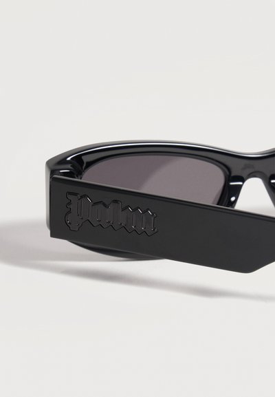 Lunettes de soleil noires avec verres foncés et branches épaisses, avec un logo "Palm" en relief en police gothique sur le côté.