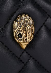 Czarna pikowana skórzana detalizacja z metaliczną złotą ozdobą w kształcie dłoni, ozdobioną kamieniami dekoracyjnymi i czarnym koralikowym okiem.