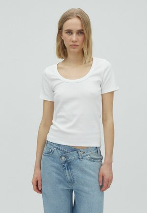 Femeie blondă purtând un tricou alb cu mâneci scurte și decolteu rotund și blugi bleu cu talie înaltă și talie desfăcută.