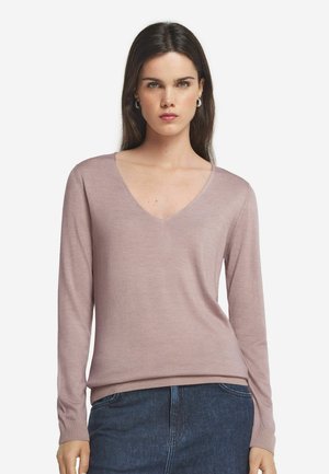 Kvinde med langt mørkt hår iført lyserød mauve V-udskåret langærmet sweater og mørkeblå jeans, stående mod en hvid baggrund.