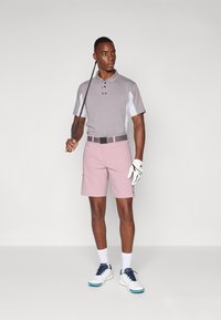 Golf outfit featuring a light gray polo with white accents, pink shorts, black belt, and white shoes with blue accents. Holding a golf club.