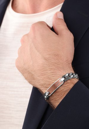 Silberfarbener Metallarmband mit Kettengliedern und einem Holzelement mit eingravierten Details, am Handgelenk getragen, teilweise von einer marineblauen Jacke bedeckt.