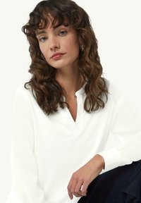 Weiße Bluse mit langen Ärmeln und V-Ausschnitt, mit gerafften Bündchen, die eine weiche Textur aufweist. Das Model posiert mit braunen, welligen Haaren vor einem neutralen Hintergrund.