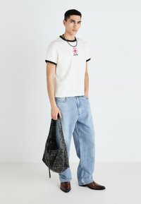 White t-shirt with black ribbed collar and red print, paired with light blue denim jeans and a textured black bag, complemented by pointed brown boots.