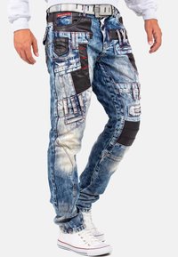 Jeans en denim bleu délavé avec des accents en cuir noir, plusieurs poches, des surpiqûres contrastantes et une coupe fuselée. Associés à des baskets blanches.