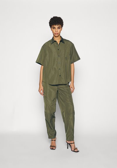 Olijfgroene, kortegesleeve shirt met een zak, gecombineerd met bijpassende tapered broek. Zachte stof met een iets gerimpelde textuur. Zwarte hakken.