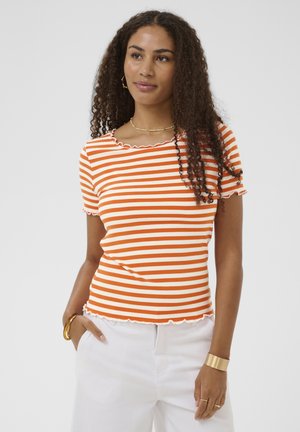 Frau mit lockigem Haar, die ein orange-weiß gestreiftes Kurzarmoberteil und weiße Hosen trägt, mit goldschmuck am Handgelenk und Hals.