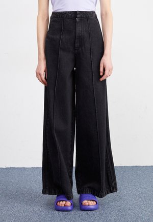 Personne portant un pantalon noir taille haute à jambes larges, associé à des sandales slides violettes à bouts ouverts, debout sur un tapis bleu.