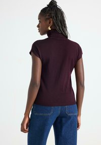 Kvinna med flätat hår, som bär guldörhängen, en kortärmad mörk vinröd polotröja och blå jeans, stående framför en vit bakgrund.