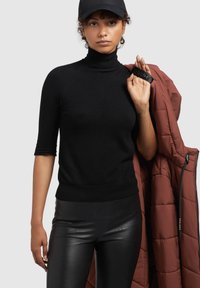 Kvinde iført sort kasket, sort turtleneck med ærmer ved albuen og sorte læderbukser, som holder en rustfarvet quiltet jakke over sin skulder.