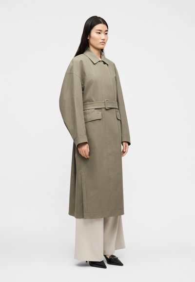 Γυναίκα που φορά ένα μακρύ, ελιτί πράσιν ο παλτό με ζώνη πάνω από φαρδιά μπεζ παντελόνια και μαύρα γόβες με μυτερή μύτη, στέκεται μπροστά σε ένα απλό λευκό φόντο.