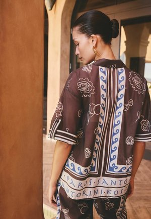 Woman with hair in a bun wearing a brown shirt and pants featuring blue and white patterns and text, standing in an arched, warm-toned corridor.