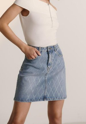 Femme portant un top blanc asymétrique et une jupe en denim bleu clair avec des coutures en forme de losange, main posée négligemment dans la poche.