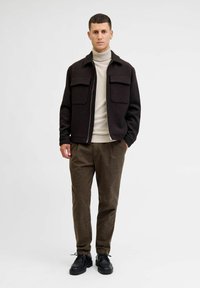Mörk ullblandad jacka med två framfickor, beige polotröja, bruna strukturerade byxor och svarta snörskor mot en neutral bakgrund.