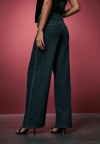 Szerokie nogawki, ciemnoniebieskie dżinsy z wysokim stanem. Ozdobne nity wzdłuż boków i na tylnych kieszeniach. Gładka tekstura oraz dopasowany krój.