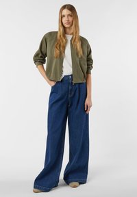 Kurtka bomberka w oliwkowej zieleni na zamek błyskawiczny, zestawiona z szerokimi jeansami o luźnych nogawkach w kolorze niebieskim. Gładka faktura, luźny krój i podstawowy biały t-shirt pod spodem.