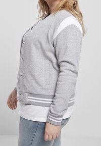 Femme portant une veste universitaire grise avec des rayures blanches sur les poignets et les épaules, par-dessus une chemise blanche et un jean bleu clair.