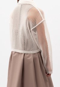Giacca corta in rete bianca con colletto rotondo, caratterizzata da una trama a intreccio aperto. Abbinata a una gonna plissettata marrone, in tessuto morbido.