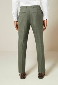Tailored green trousers with a textured finish, two back pockets with buttons, and a straight-leg cut. Worn with brown leather shoes.