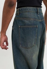 Sötétkék denim farmernadrág laza szabással, látható fakulással és két hátsó zsebbel. Magas derékkal és texturált anyaggal rendelkezik.