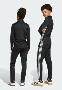 Svart träningsdräkt med zip-up-jacka och tajta byxor, med tre vita ränder på sidorna och en slät, texturerad tyg.