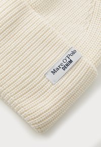 Bijela pletena kapa napravljena od teksturirane pređe s rebrastim dizajnom. Ima pravokutnu oznaku s natpisom "Marc O'Polo DENIM" u plavoj i bijeloj boji.