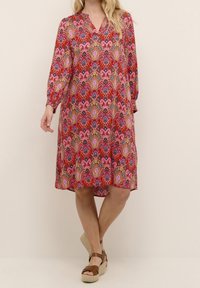 Kvinna i en knälång röd klänning med rosa, guld och blå paisleymönster och bruna kilklackar, stående med korsade ben.