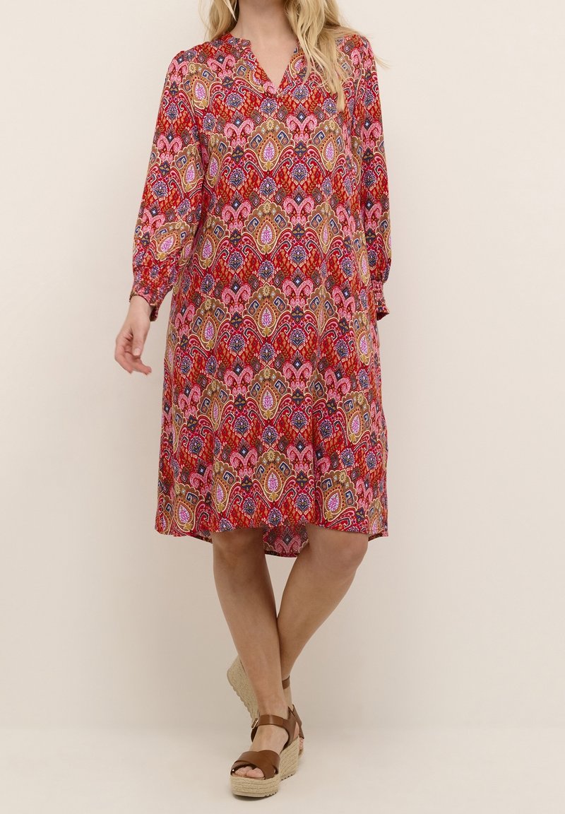 Kvinna i en knälång röd klänning med rosa, guld och blå paisleymönster och bruna kilklackar, stående med korsade ben.