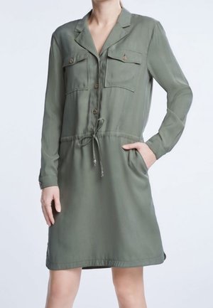 Vrouw draagt een olijfgroene shirtjurk tot op de knie met lange mouwen, borstzakken met knopen en een taille met trekkoord.