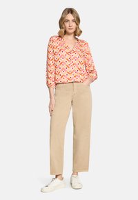 Femme debout, une main dans la poche, portant un pantalon beige, des baskets blanches et un chemisier à manches longues à motifs avec des formes rouges, orange et roses.