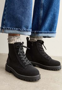 Bottines en suède noir avec un laçage à l'avant, une fermeture éclair latérale, une semelle en caoutchouc épaisse et des détails de coutures blanches, associées à des chaussettes en tricot grises et un jean.