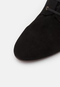 Scarpa in suede nera con punta arrotondata e superficie texturizzata, con sottili lacci neri e un bordo della suola marrone liscio.