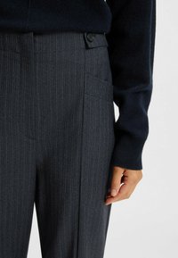 Grau gestreifte Hose aus einem strukturierten Stoff, mit einer Vorderasche und Knopfverschluss, kombiniert mit einem marineblauen Pullover.