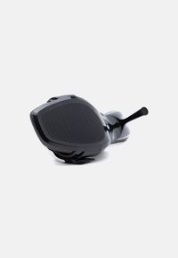 Μαύρο παπούτσι με ψηλό τακούνι από δέρμα βινυλίου, ξαπλωμένο στο πλάι, που δείχνει σόλα με αυλακώσεις και λεπτό τακούνι σε λευκό φόντο.