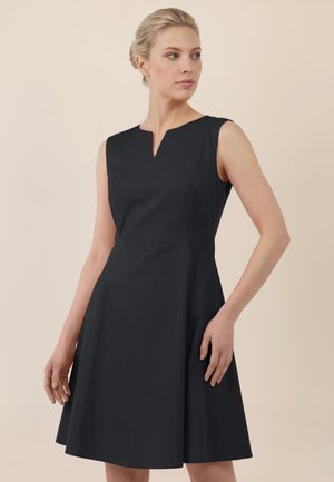 Frau mit blondem Haar in einem niedrigen Dutt, die ein ärmelloses schwarzes knielanges Kleid mit kleinem Ausschnitt vorne trägt.