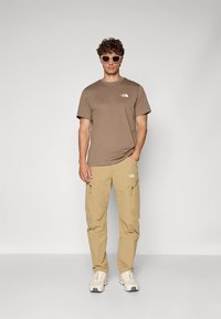 Pruun puuvillane T-särk logoga, kombineeritud beežide lahtiste pükstega, millel on taskud, ning valgete spordijalatsitega. Aksessuaarideks on päikeseprillid.