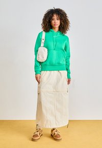 Green hoodie with front pocket, cream utility skirt with side zippers, beige sneakers with brown accents, and a small cream crossbody bag.