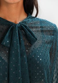 Blouse en voile teal avec un col à nœud, présentant un motif texturé de petits points réfléchissants. Tissu léger et élégant avec une finition lisse.