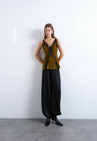 Femme debout contre un mur blanc, portant un caraco moutarde bordé de dentelle, un pantalon large noir et des bottes à talons noires.