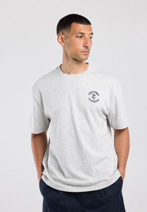 Man met kort haar, die een lichtgrijs New York Club House T-shirt en een donkere broek draagt, staat met de handen in de zakken en kijkt naar de zijkant.